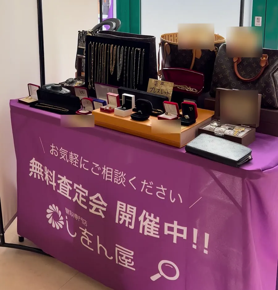 無料査定会のブース。紫のテーブルクロスに「無料査定会 開催中!!」としをん屋の文字。テーブルに見本の品物を置いている
